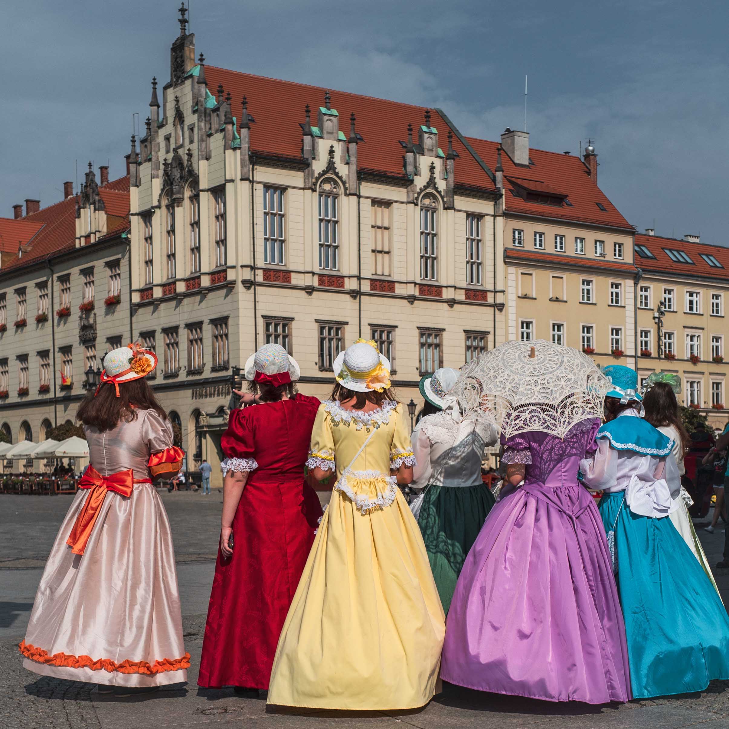 sechs Frauen mit tollem mittelalterlichen Kleid sind von hinten zu sehen und schauen in Richtung eines städtischen Gebäudes
