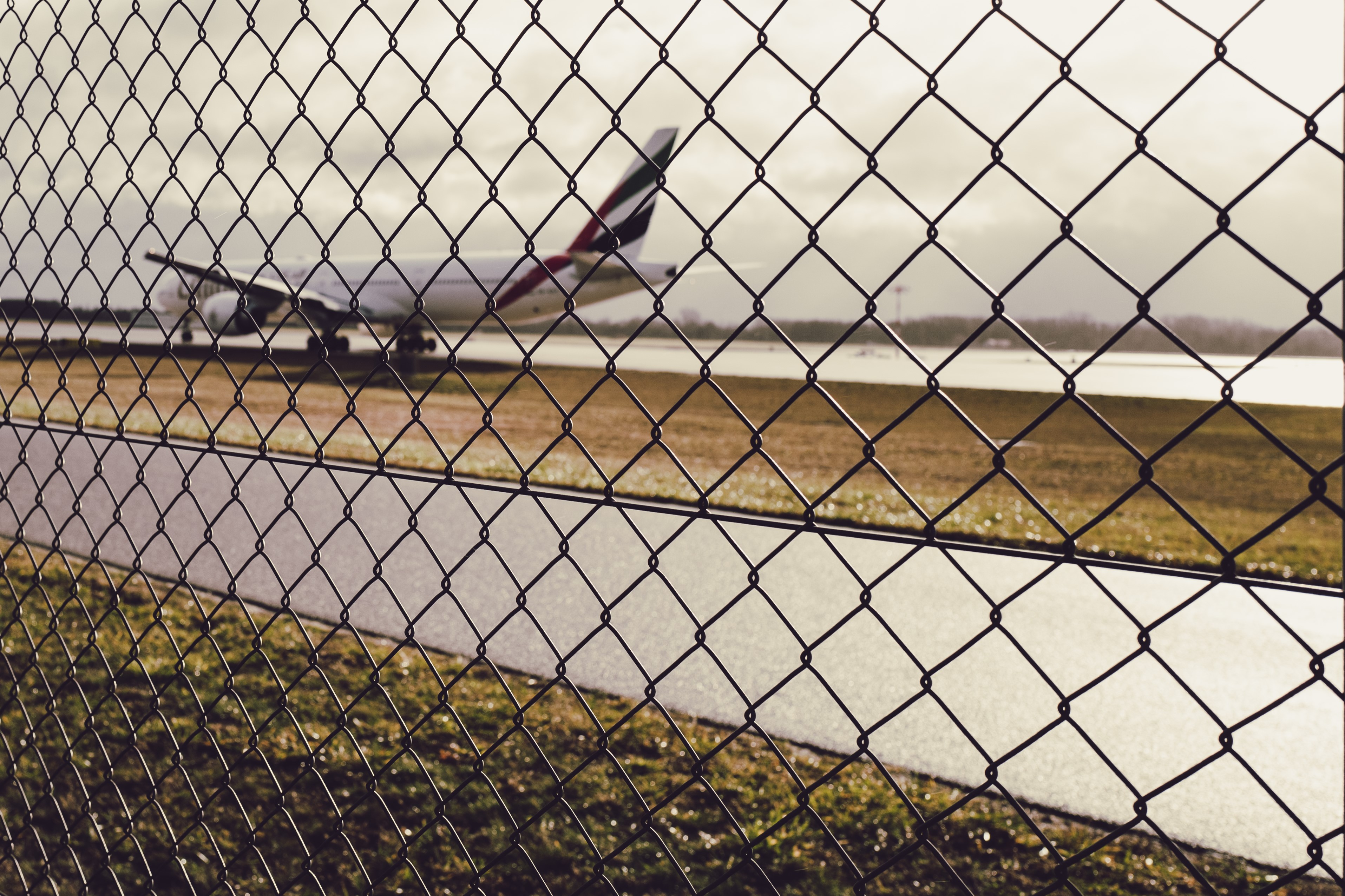 Das Bild zeigt einen Maschendrahtzaun im Vordergrund, der scharf im Fokus steht. Im Hintergrund ist ein unscharfes Flugzeug auf einem Rollfeld eines Flughafens zu erkennen. Die Umgebung wirkt trüb und bewölkt mit einer diffusen Beleuchtung.