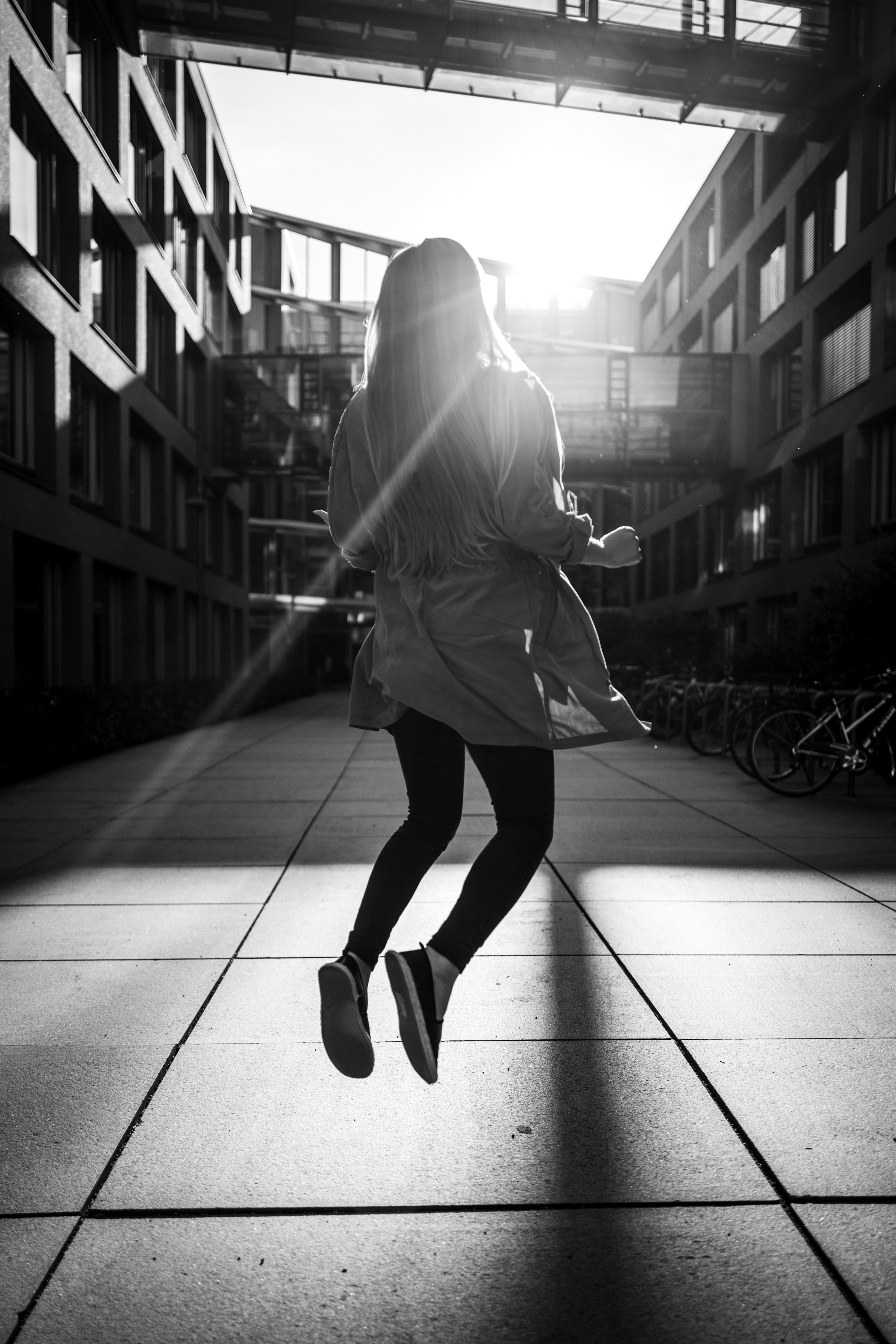 Das Bild zeigt eine schwarz-weiße Fotografie. In der Aufnahme springt eine Person zwischen Schatten und Licht. Die Umgebung ist urban, mit modernen Gebäuden auf beiden Seiten und einem Gehweg in der Mitte. Sonnenlicht durchflutet die Gebäude und erzeugt kontrastierende Schatten auf dem  Boden.