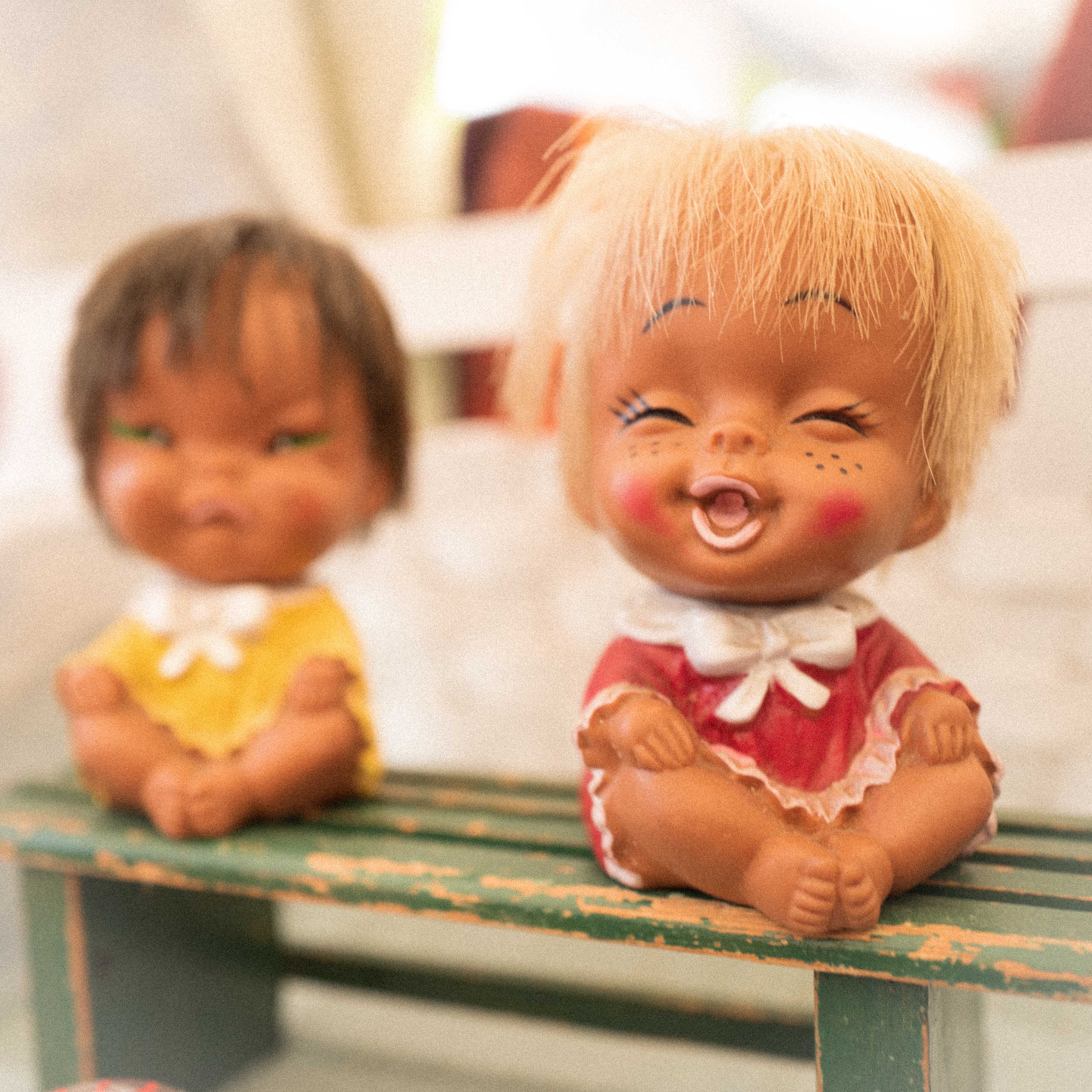 Das Bild zeigt zwei kleine Figuren, die auf einer grünen, verwitterten Bank sitzen. Eine Figur trägt ein gelbes Outfit und die andere ist in Rot mit weißen Details gekleidet. Eine von den Puppen hat ein lachendes Gesicht, diese ist scharf zu sehen, die andere schmolllt und ist unscharf zu sehen. Die Puppen sollen zwei Kinder darstellen.