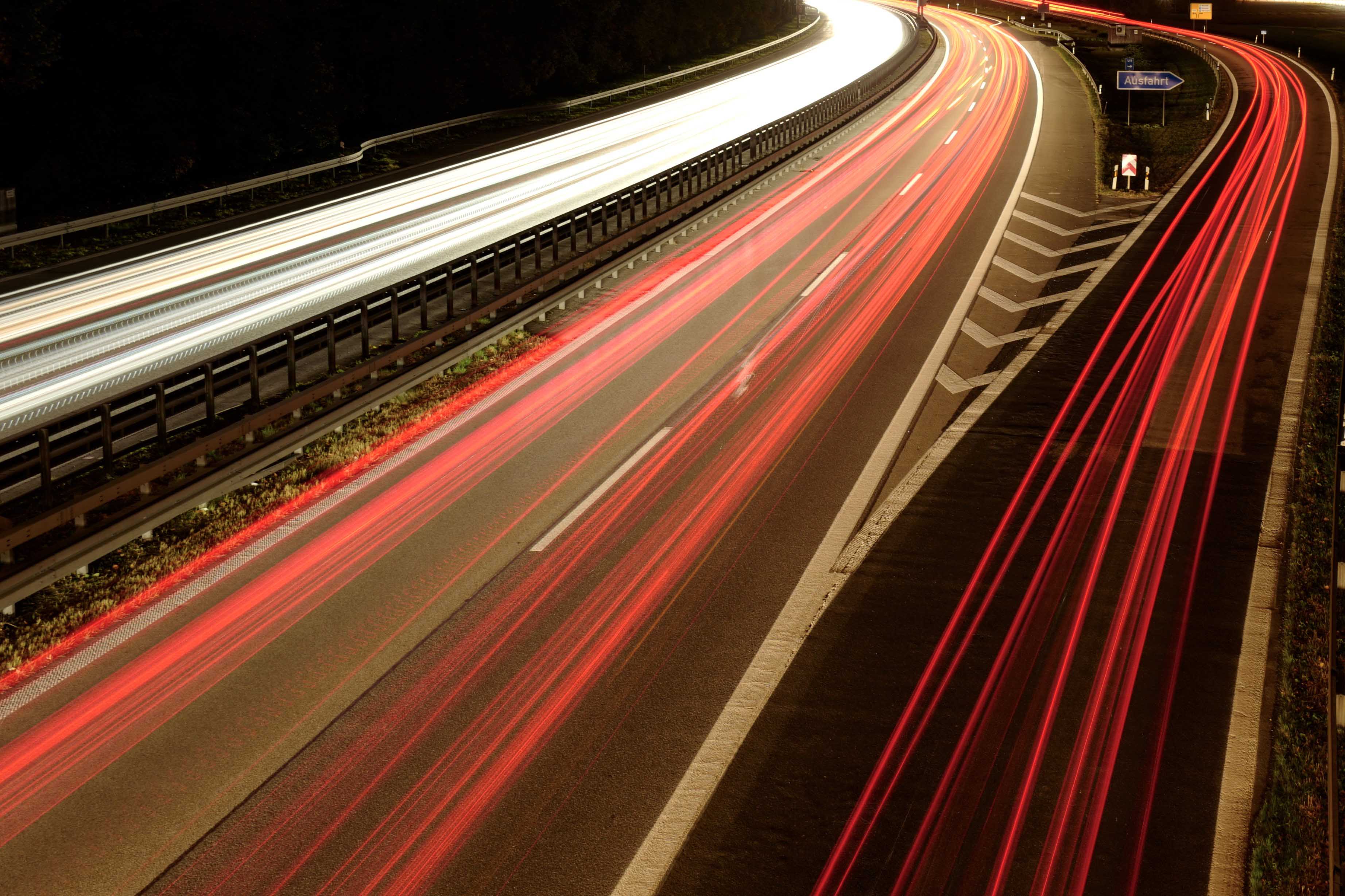 Das Bild zeigt eine belebte Autobahn bei Nacht. Die roten Rücklichtern der Autos, sind auf der rechten Seite des Bildes zu sehen. Weiße Lichtspuren, die Scheinwerfern der Autos, sind auf der linken Seite des Bildes sichtbar. Die Kurvenform der Straße und die Lichtspuren erzeugen ein dynamisches und kinetisches Gefühl im Bild. Hochauflösene Aufnahme von Simon Fritz mit seiner Leica Kamera.