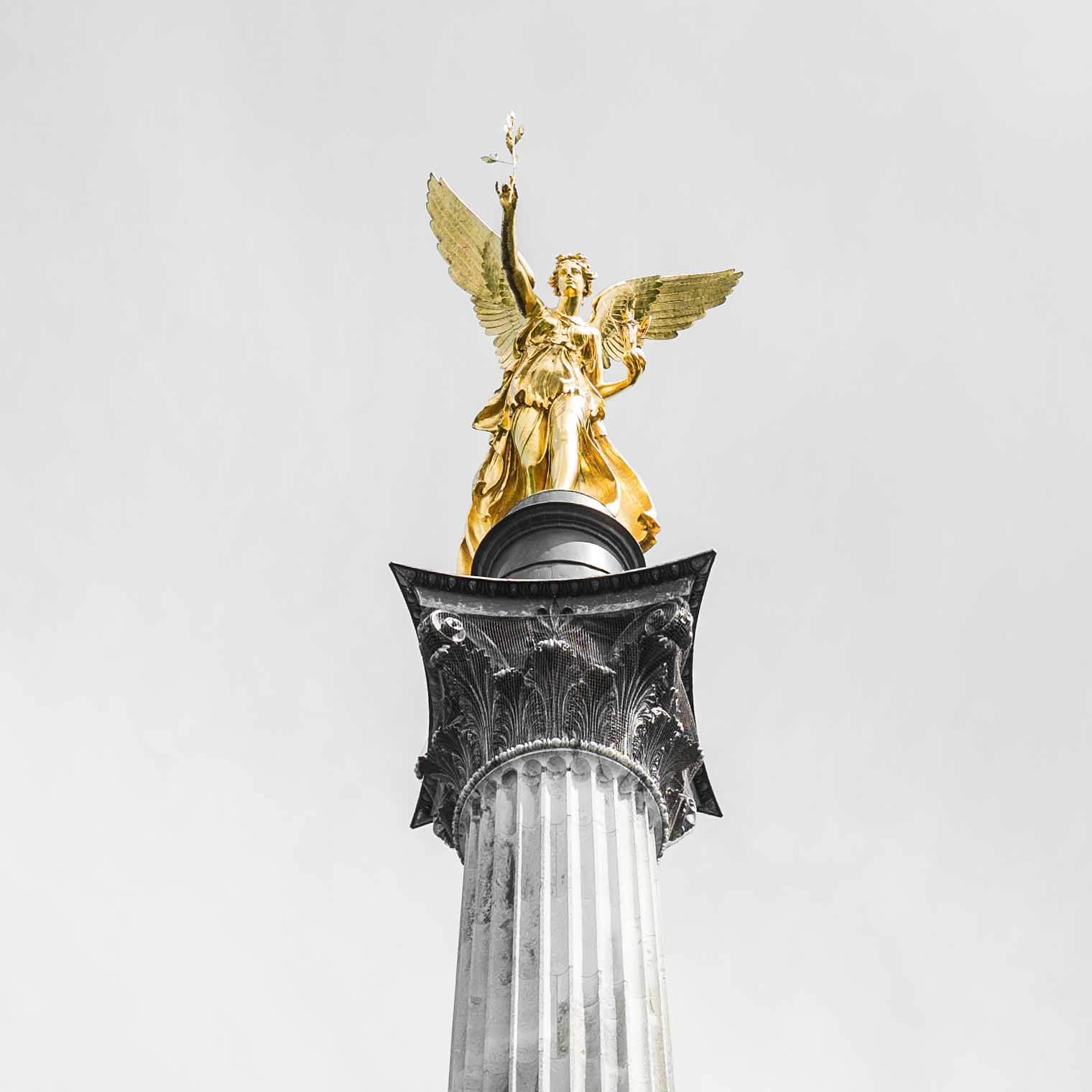 Zu sehen ist die der obere Teil des Friedensengels in München. Die Statue stellt eine Figur mit Flügeln dar, die einen Lorbeerkranz in der Hand hält. Der Himmel im Hintergrund ist bewölkt und links und rechts sind grüne Baumkronen zu sehen.
