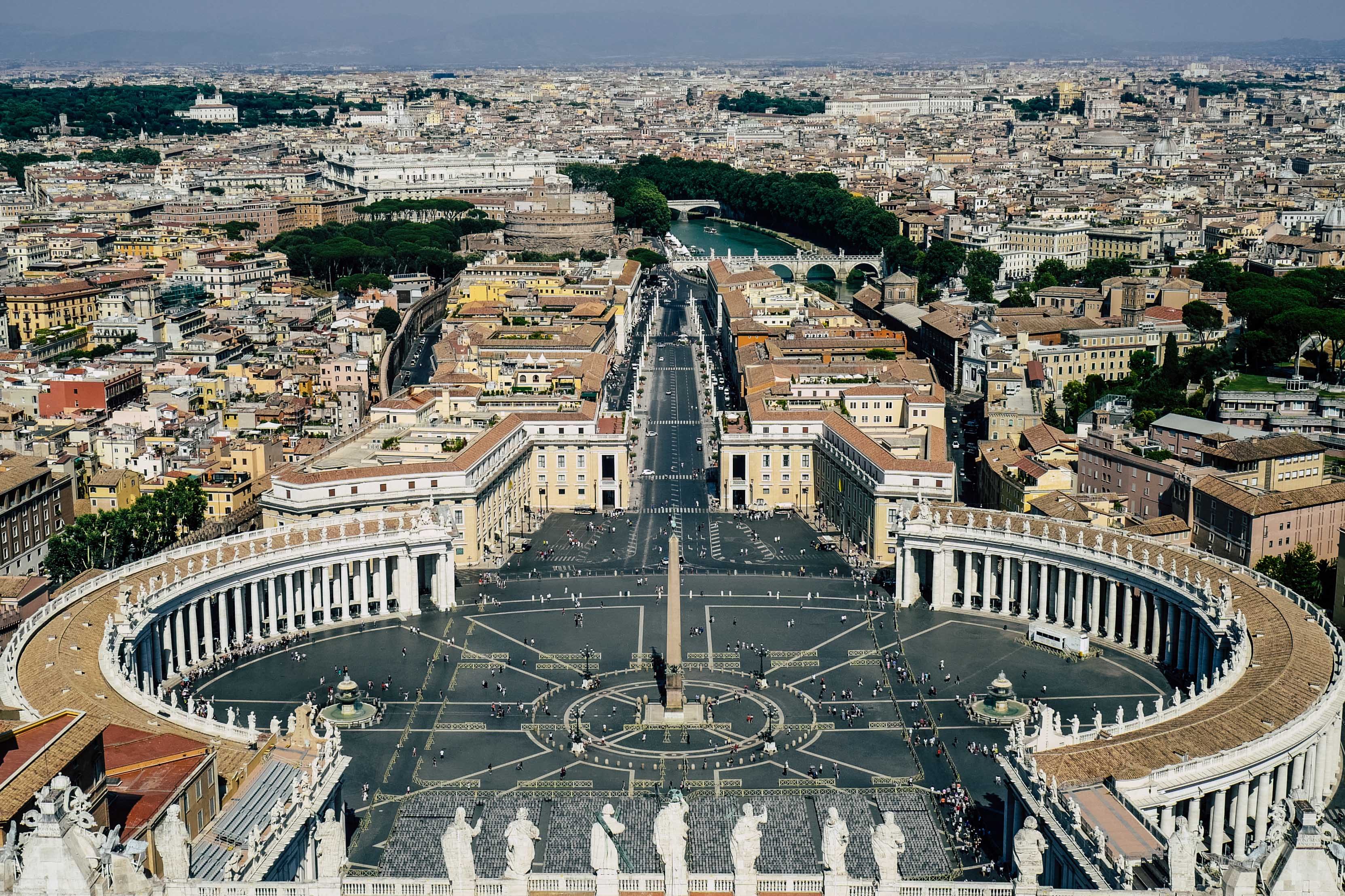 Das Bild zeigt den Petersplatz in Rom, umgeben von der beeindruckenden Architektur der Vatikanstadt und der Stadt Rom im Hintergrund. Der Platz ist prominent dargestellt, mit seiner ikonischen ovalen Form, umgeben von einer Kolonnade. In der Mitte des Platzes steht ein Obelisk. Die Straßen führen zum Platz sind gut organisiert und symmetrisch. Die Architektur des Vatikans ist weiß und majestätisch, mit Statuen, die die Oberseite der Kolonnaden schmücken. Im Hintergrund sieht man die dichte Bebauung Roms mit seinen charakteristischen gelben und braunen Gebäuden. Der Fluss Tiber schlängelt sich durch die Stadt im Hintergrund. Aufgenommen vom Dach des Petersdoms. Ein Perfektes Wandbild für ein italienisches Restaurant oder ein Wohnzimmer.