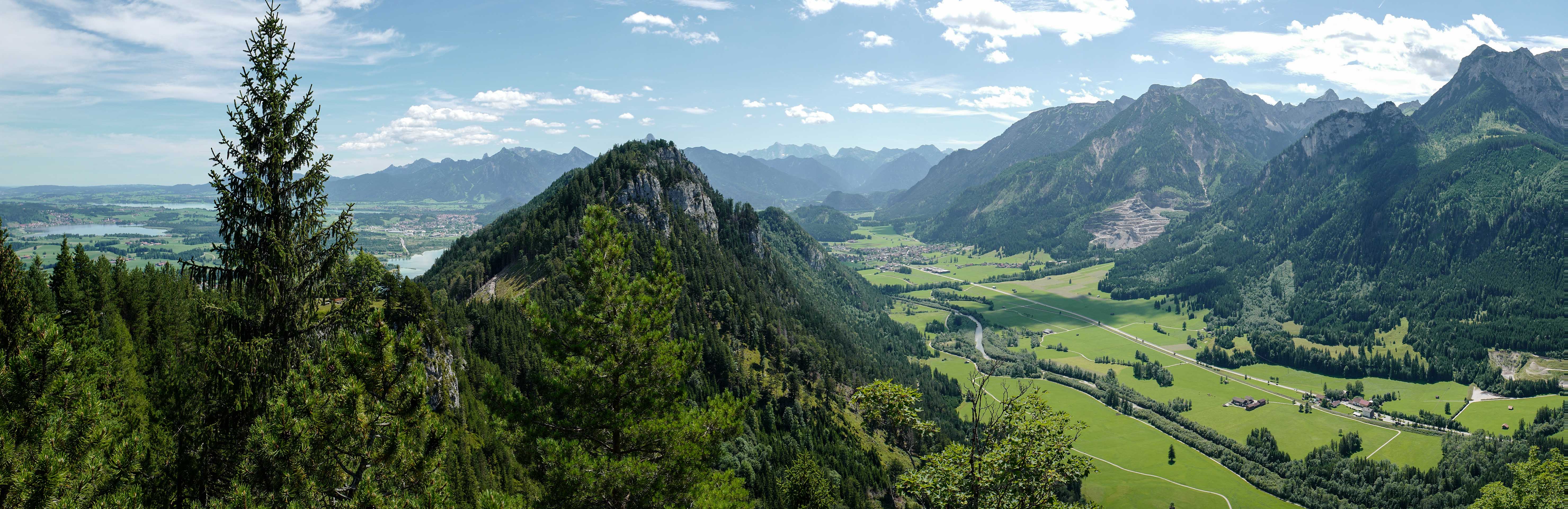 Das Bild zeigt ein atemberaubendes Panorama der Allgäuer Alpen, das von üppigen grünen Tälern, schroffen Berggipfeln und einem klaren Himmel geprägt ist. Im Vordergrund befindet sich ein felsiger Berg, der mit grünen Bäumen und Vegetation bedeckt ist. Jenseits dieses Berges erstrecken sich ausgedehnte grüne Täler, die von Wäldern und offenen Flächen durchsetzt sind. Ein gewundener Fluss schlängelt sich durch das Tal. In der Ferne erheben sich mehrere Berggipfel majestätisch gegen den Horizont; ihre schroffen Umrisse erzeugen einen dramatischen Kontrast zum Himmel. Diese Sommerlandschaft wurde vom heidelberger Fotografen Simon Fritz aufgenommen und ist in seinem Wandbilder Shop erhältlich.