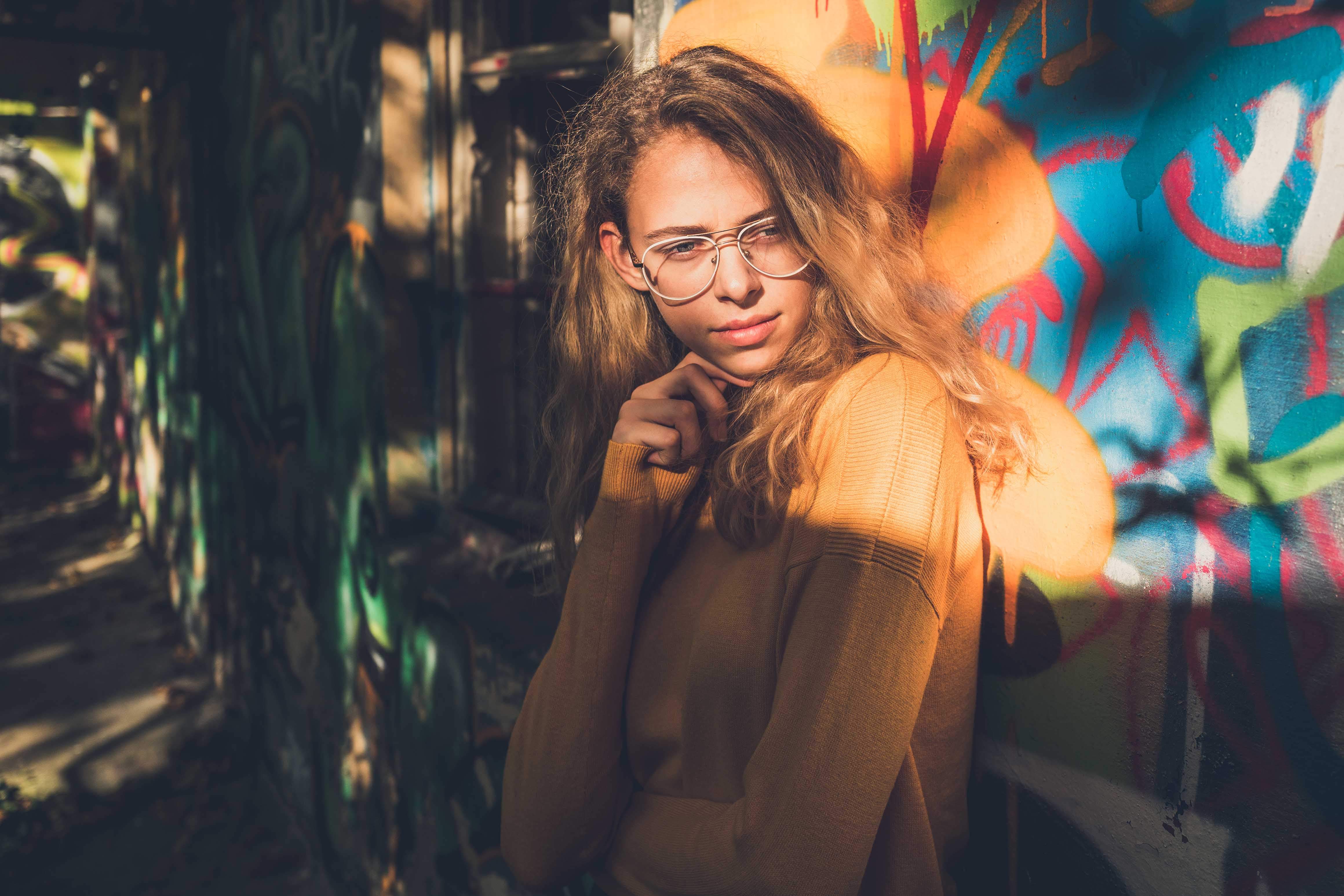 Portraitaufnahme einer Frau, die vor einer farbenfrohen Graffiti-Wand steht. Die Person trägt einen langärmeligen orangen Pullover. Die Graffiti im Hintergrund sind lebhaft und zeigen verschiedene Farben und abstrakte Formen. Es scheint, dass das Sonnenlicht von einer Seite auf die Person scheint, was einen Kontrast von Licht und Schatten erzeugt.