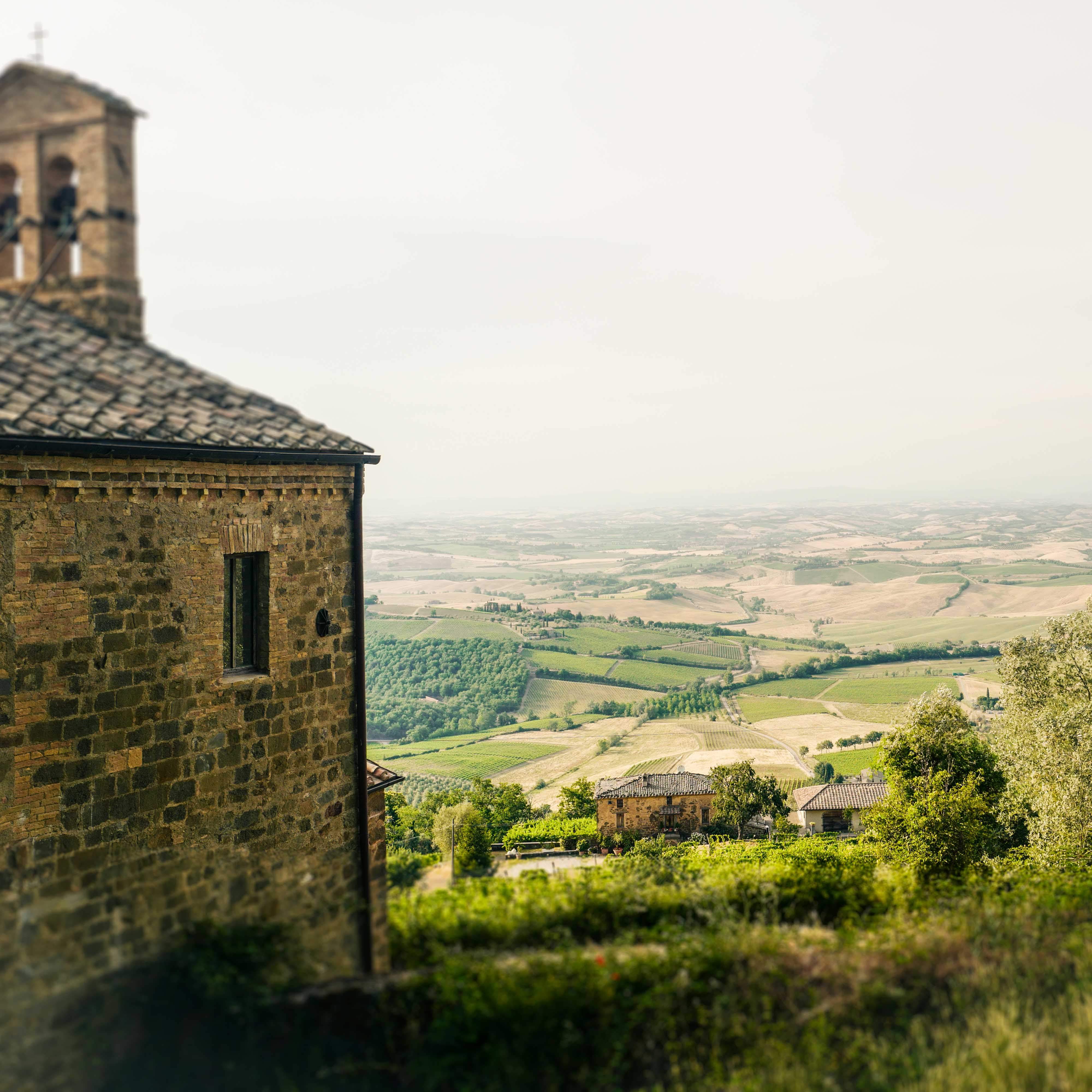 Das Bild zeigt eine malerische Aussicht auf die Landschaft der Toskana. Im Vordergrund ist ein Teil eines alten, steinernen Gebäudes mit einem sichtbaren Fenster und Dach zu sehen. Die Landschaft ist geprägt von sanften Hügeln, grünen Feldern und Waldstücken, die über das Land verstreut sind und auf ländliche Besiedlung hinweisen.