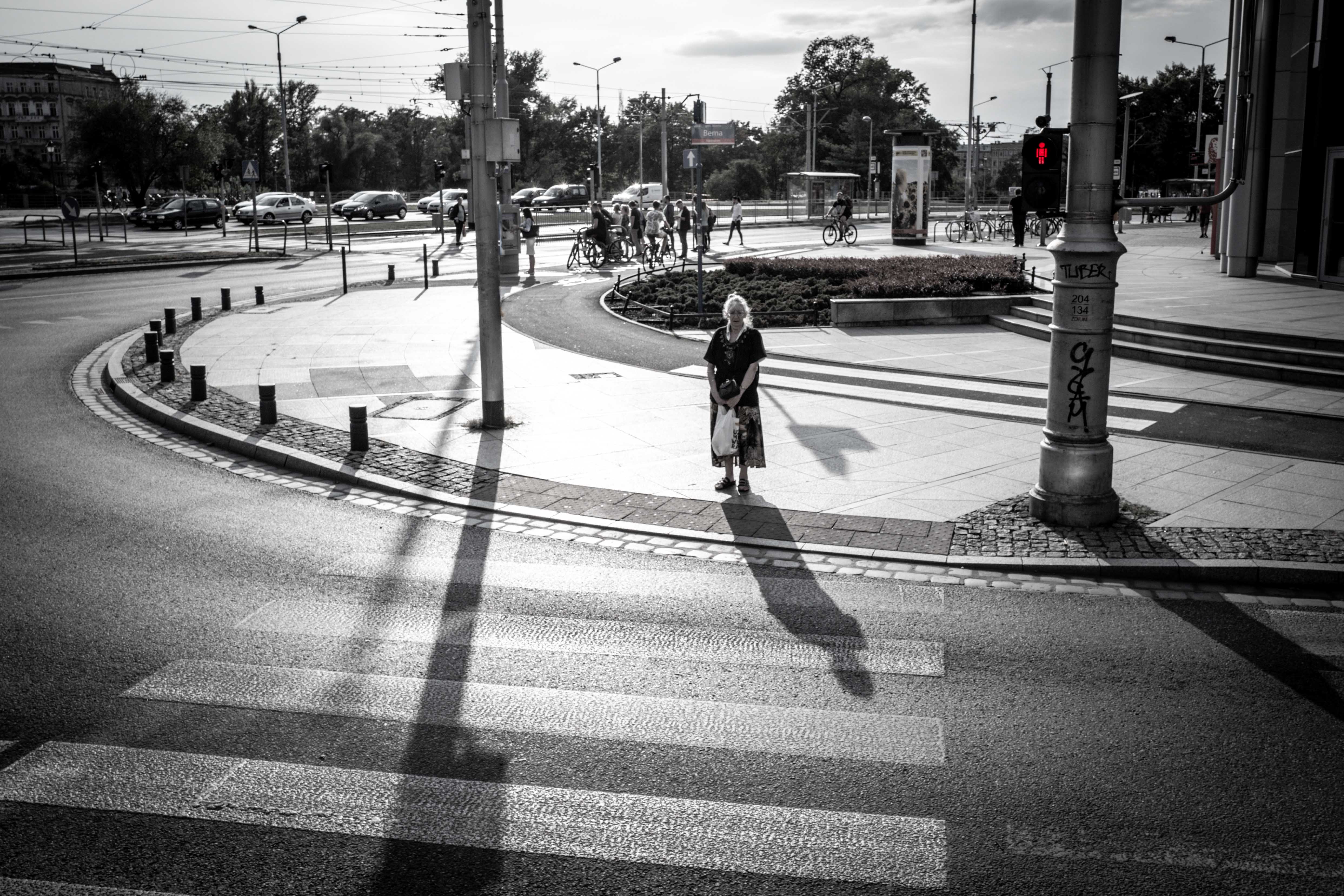Ein schwarzweiss Bild, aufgenommen mit einer Leica Kamera in Breslau. Das Bild zeigt eine städtische Umgebung mit einer Straße, einem Gehweg und Verkehrszeichen. Eine Person steht auf dem Gehweg nahe der Straßenkreuzung und wartet darauf, die Straße zu überqueren. Die Ampel zeigt ein rotes Licht an. Im Hintergrund sind weitere Personen sichtbar, die sich auf dem gegenüberliegenden Gehweg aufhalten. Die Sonne scheint hell und erzeugt deutliche Schatten der Objekte auf der Straße und dem Gehweg. Mit Klick auf das Bild, kann man das Motiv als Wandbild erwerben.