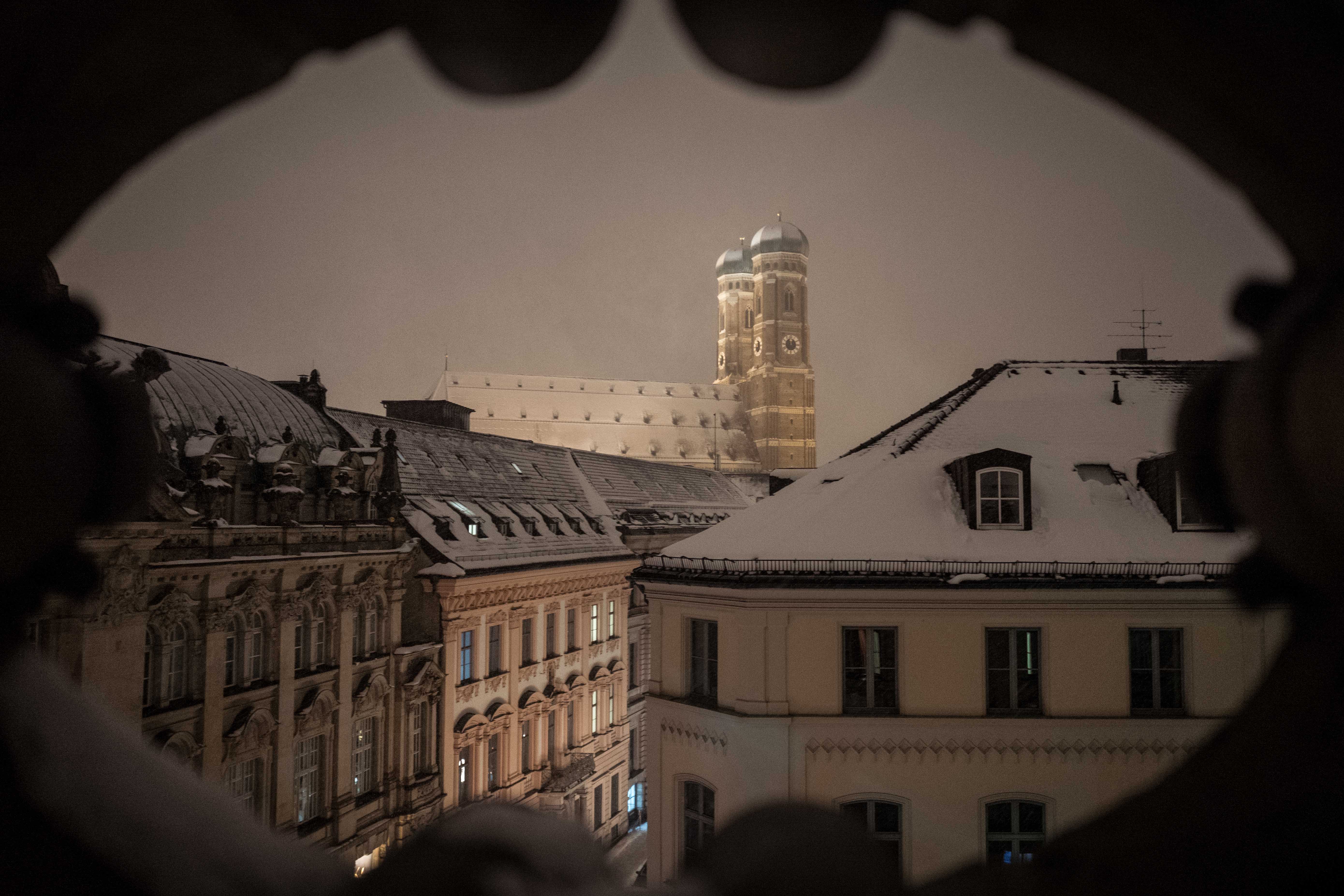 Das Bild zeigt eine nächtliche, schneebedeckte Stadtlandschaft mit historischen Gebäuden und der Frauenkirche München im Hintergrund. Die Umgebung ist von Schnee bedeckt und gibt ein ruhiges, friedliches Gefühl. Im Vordergrund sind schneebedeckte Dächer von Gebäuden zu sehen, die architektonische Details aufweisen. Es gibt eine Reihe von Fenstern an den Gebäuden im Vordergrund, einige sind beleuchtet. Der Hintergrund wird von der beleuchteten Frauenkirche dominiert; Die Aufnahme scheint durch eine Art Rahmen oder Öffnung gemacht worden zu sein, da die oberen Ränder des Bildes unregelmäßig geformt sind. Aufgenommen mit der Leica Q, als Wandbild zu erwerben.