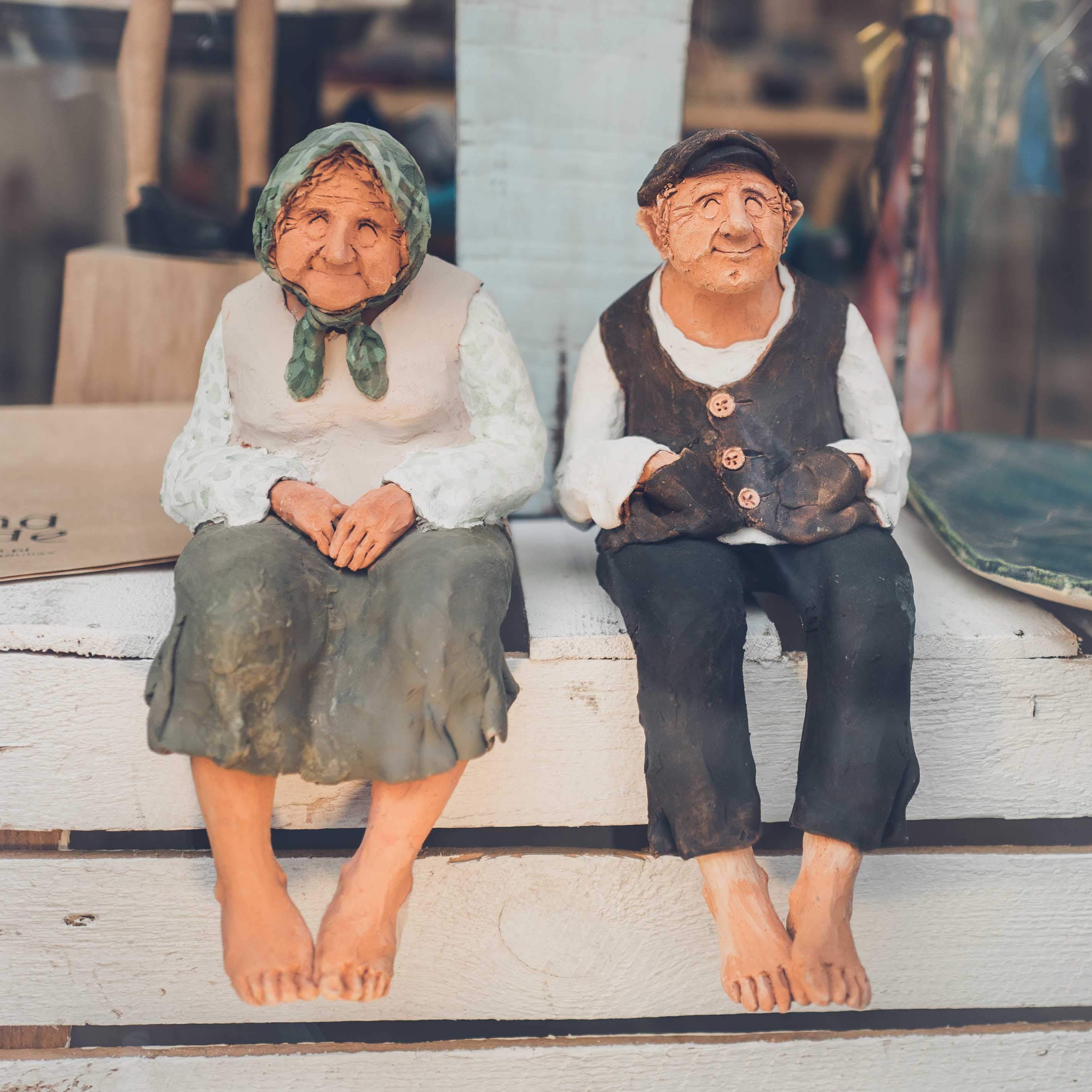 Zu sehen ist eine Fotografie von zwei Ton-Figuren von einem älteren Ehepaar welche Barfuß und mit einem Lächeln auf den Lippen dasitzen. Ein süßes Wandbild welches als Kleinformat im Wandbilder- Shop von Simon Fritz zu erwerbenkaufen gibt.