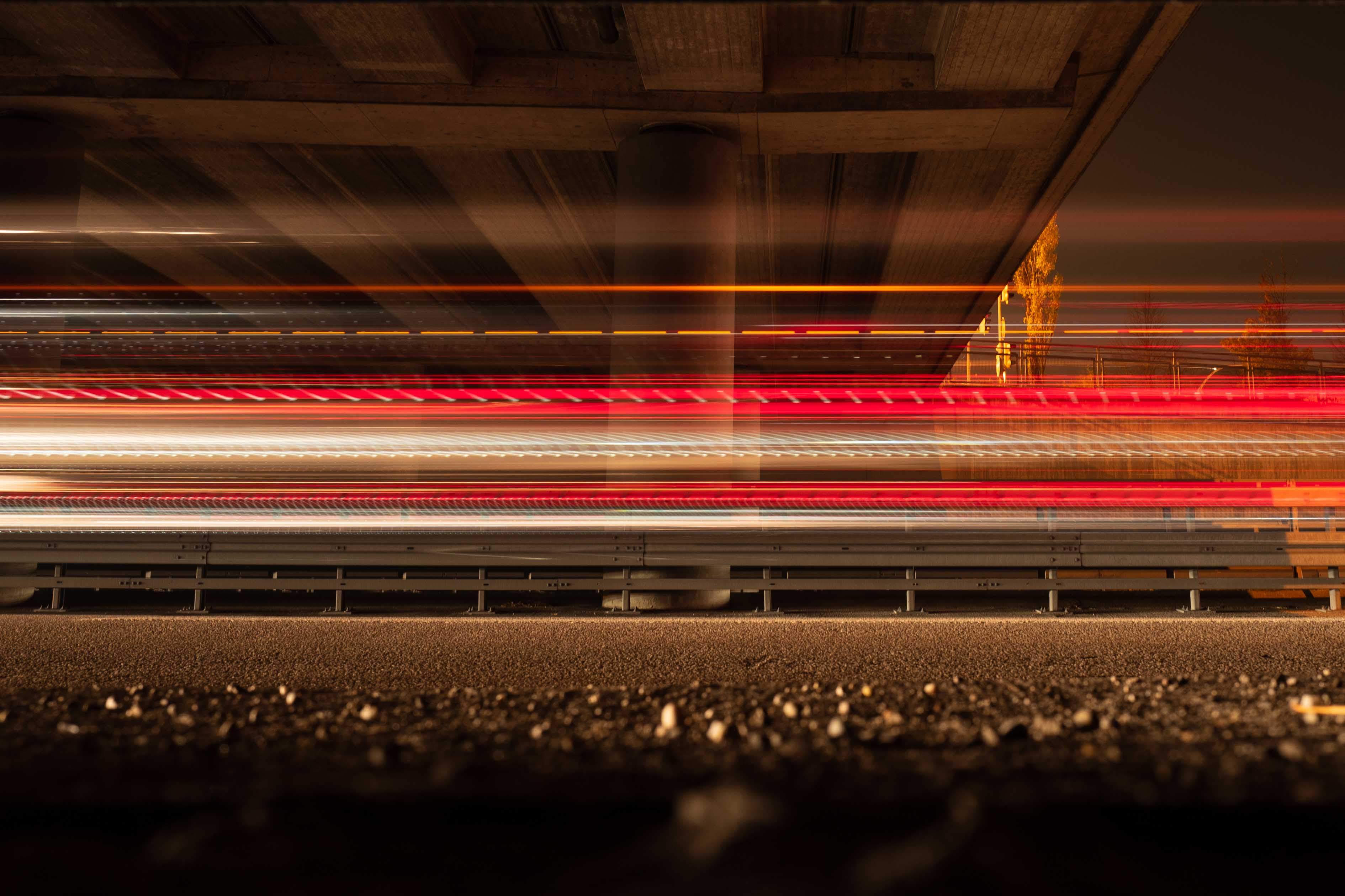 Das Bild zeigt eine Langzeitbelichtung von Fahrzeuglichtern, die unter einer Autobahnbrücke hindurchfahren. Die Lichtstreifen sind rot und weiß, was auf Rück- und Scheinwerfer der Fahrzeuge hinweist. Die Unterseite der Brücke ist sichtbar und wird von den Lichtern beleuchtet. Die Atmosphäre des Bildes ist dynamisch und dramatisch durch die Bewegung der Lichter. Fotografiert von Simon Fritz und seiner Leica Kamera bei Nacht.