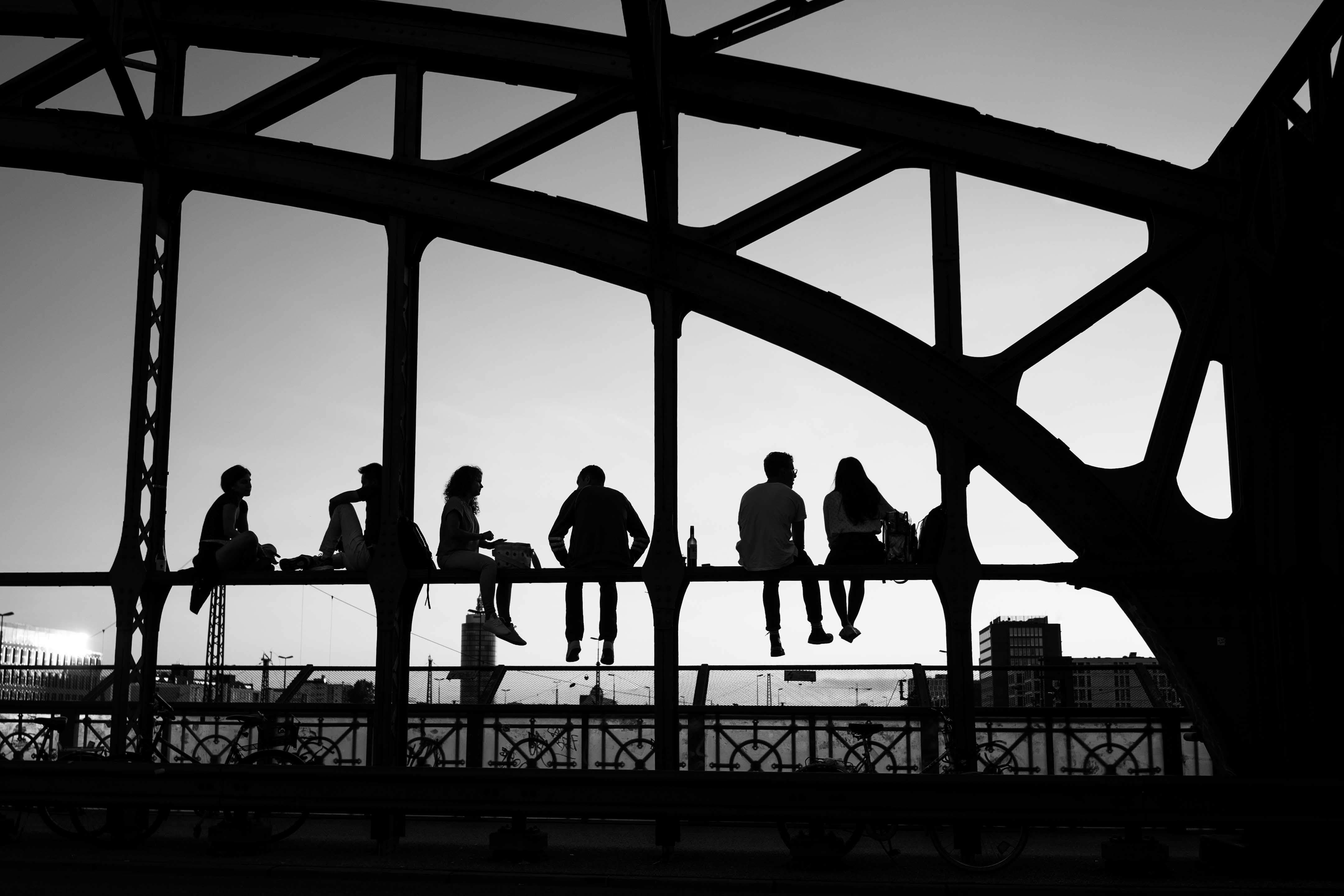 Das Bild zeigt fünf Personen, die auf einer Erhöhung der Hackerbrücke München sitzen und den Blick in die Ferne richten. Die Personen sind als Silhouetten dargestellt und scheinen in ein Gespräch vertieft oder genießen einfach den Ausblick. Die Struktur der Brücke ist geometrisch und bildet ein interessantes Muster im Hintergrund. Das Bild ist in Schwarzweiß und zeigt eine Szene bei Sonnenuntergang. Im Hintergrund ist der klare Himmel sichtbar, der von Gebäuden am Horizont unterbrochen wird. Das Motiv kann man als Wandbild erwerben.