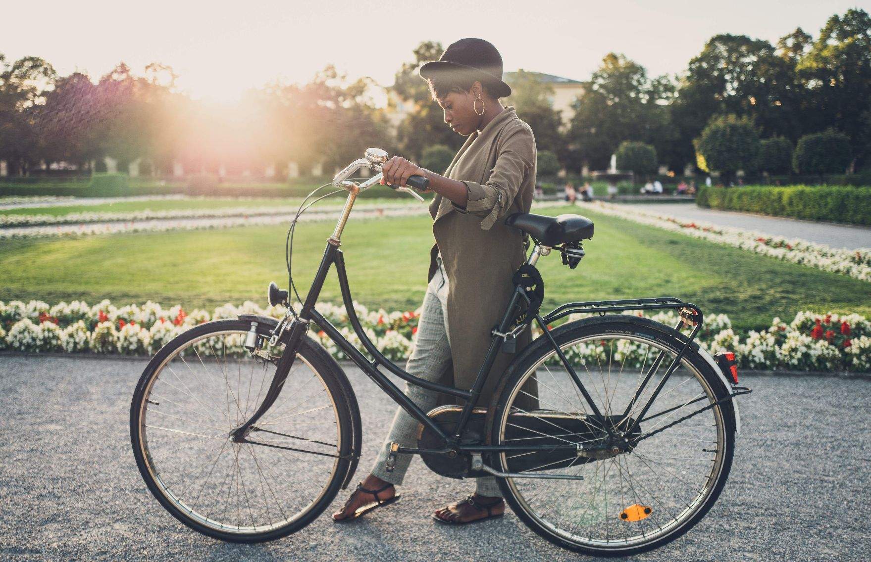 Das Bild zeigt eine Person, die ein klassisches, schwarzes Fahrrad im Hofgarten in München hält. Die Person trägt einen Hut, einen langen Mantel, sowie offene Schuhe. Der Hintergrund zeigt die gepflegte Anlage mit grünem Rasen und blühenden Blumen unter einem klaren Himmel. Sonnenlicht durchflutet die Portraitaufnahme und erzeugt eine warme und einladende Atmosphäre. Aufgenommen vom Heidelberger Fotograf Simon Fritz.